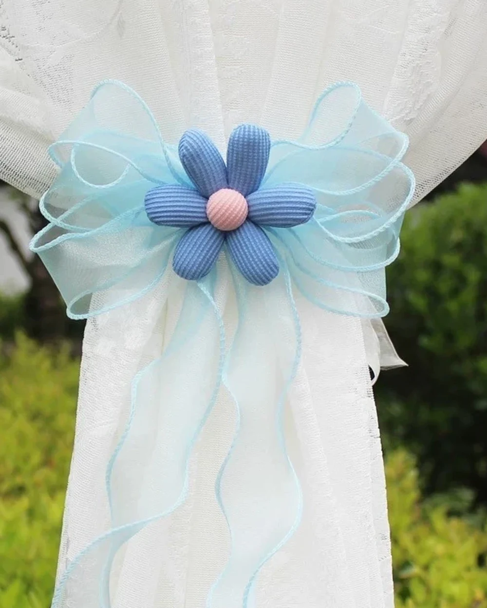 Blue six-petal fabric flower with a pink centre and pale aqua sheer ribbon bow on a white curtain.