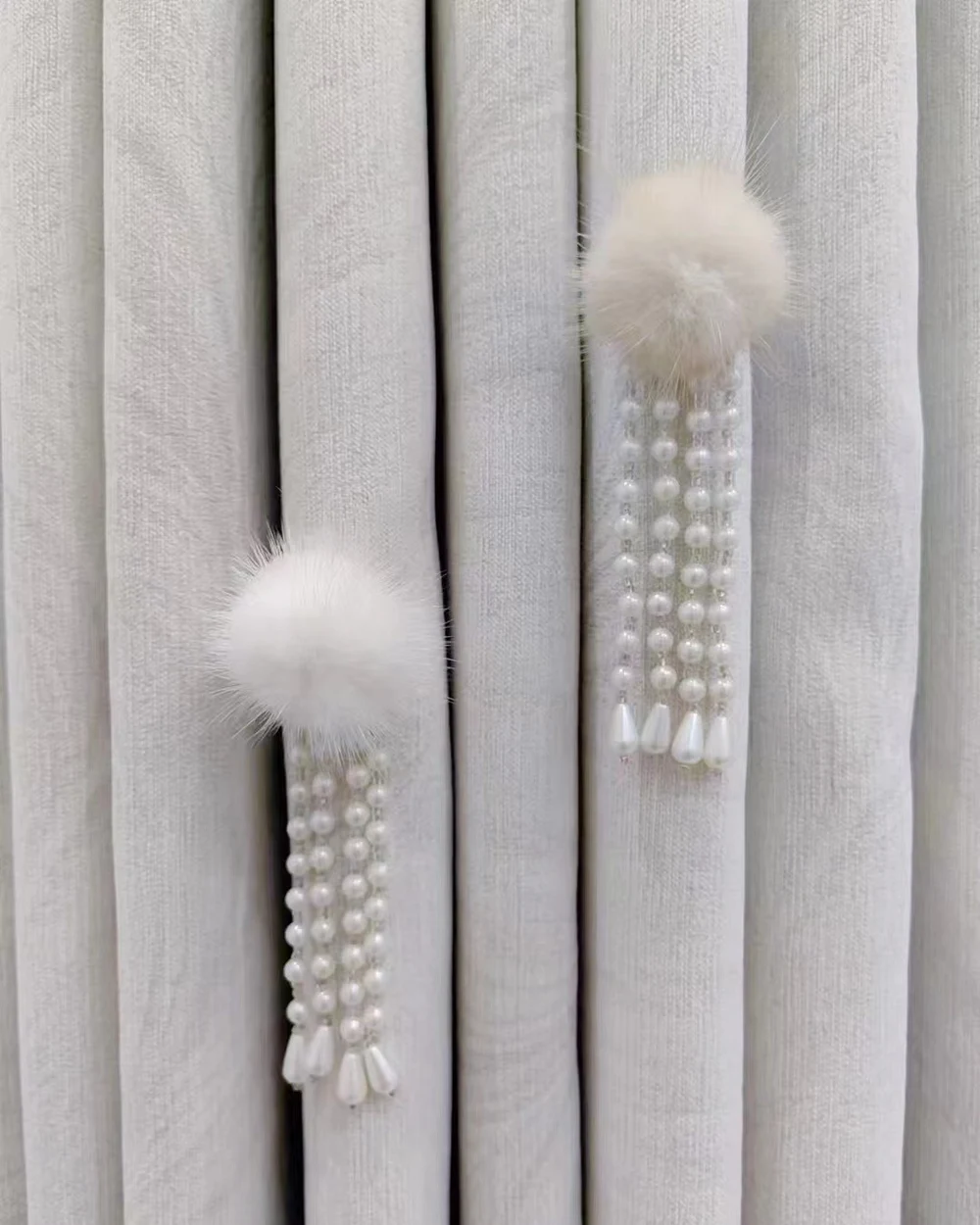 Two soft ivory pom-pom curtain ornaments with falling pearl-look bead strands on pale grey curtains.