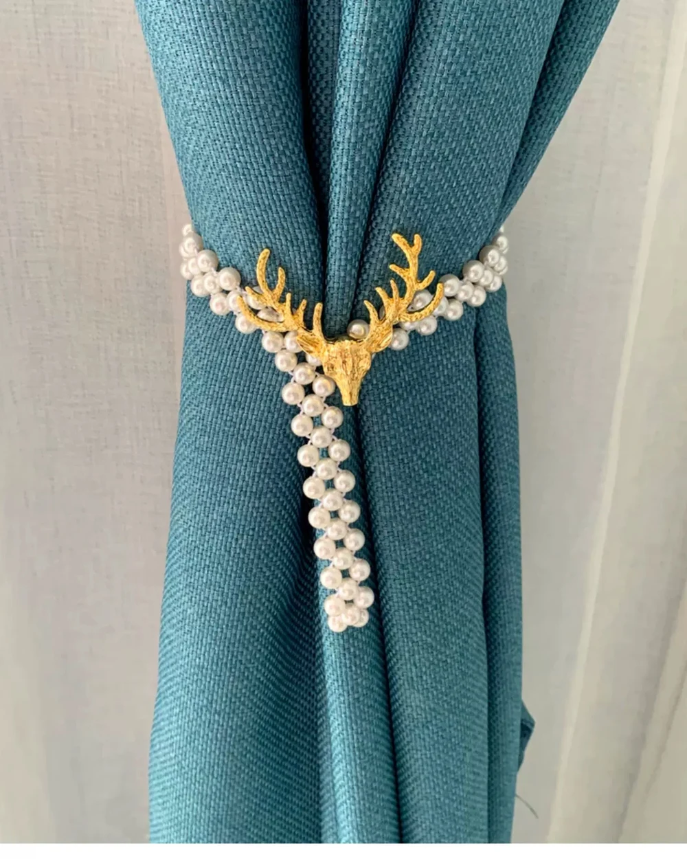 Gold-tone stag head over layered pearl-look strands gathering a deep teal curtain.