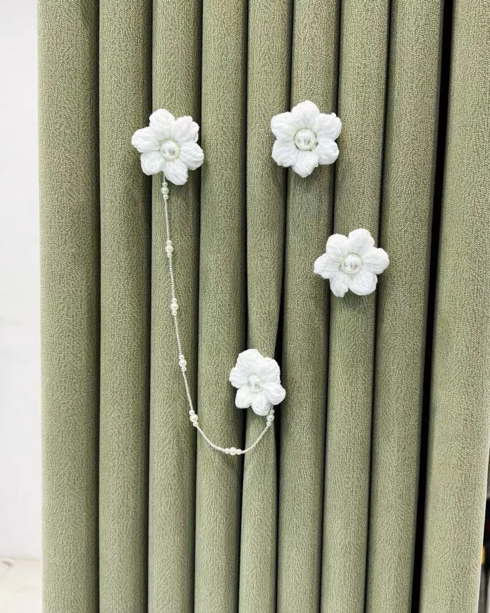 Four ivory fabric blossoms linked by a fine pearl-look chain against a sage curtain.