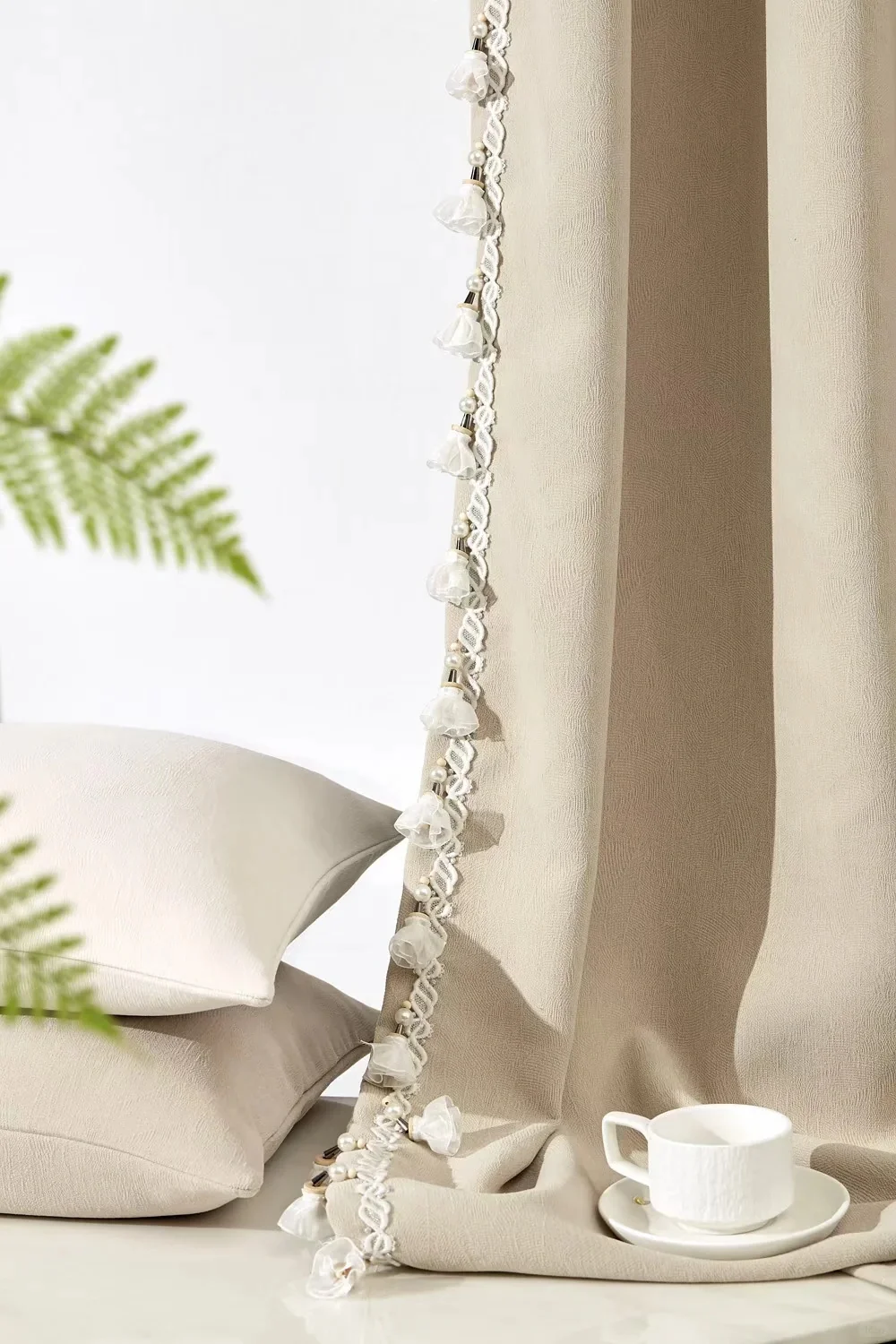 Cream curtain edge with dangling floral/leaf trim beside a pale chair and fern.