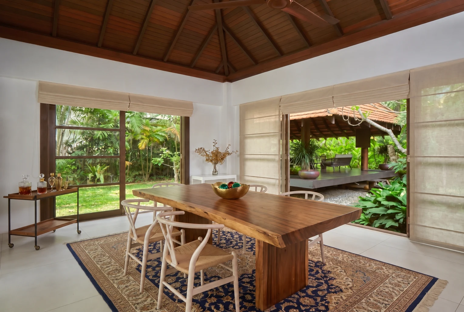 Cream Roman-style fabric blinds frame garden-facing dining-room openings beneath a timber ceiling, with a wooden table and views of tropical greenery.