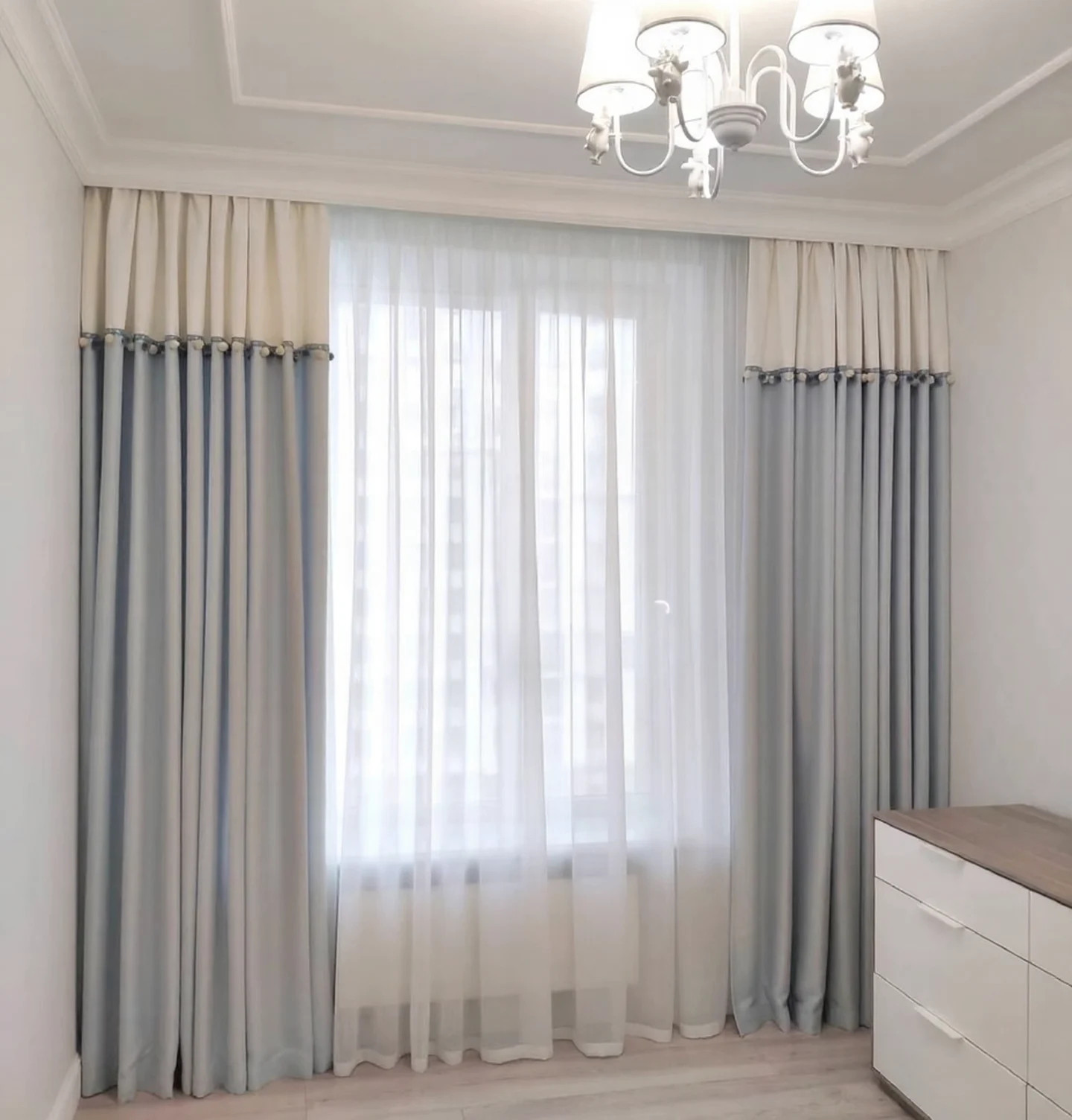 Curtain pair with ivory top fifth and dusty powder-blue long lower panels, narrow braided join carrying small alternating ivory and blue pom-poms, white full-height sheers behind. — supplied curtain design reference.