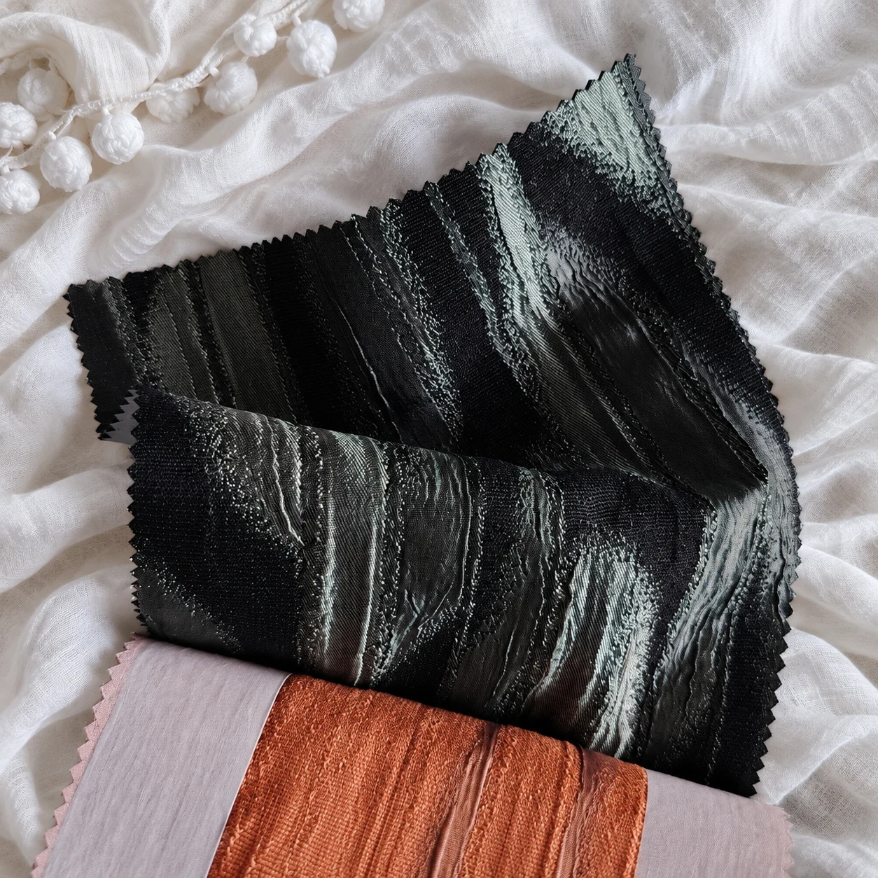 Black fabric with irregular silvery streaks over pale lilac and terracotta samples, with white pom-pom sheer.