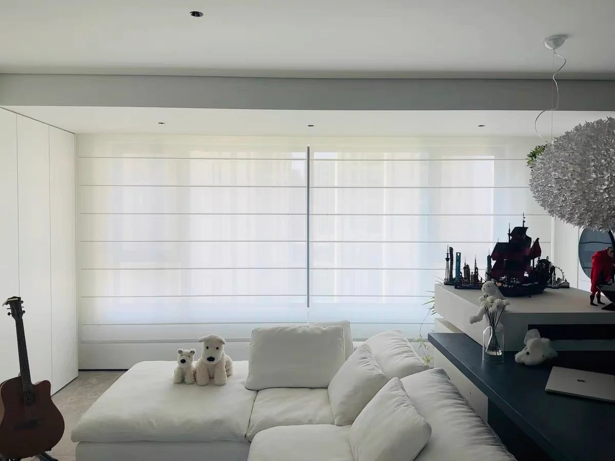 Two broad white flat Roman blinds with horizontal batten seams cover a living-room window above a cream sofa.